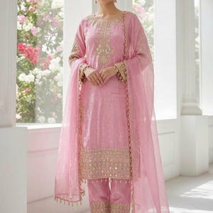 May include: A light pink salwar kameez ensemble with gold embroidery and mirror work. The outfit includes a tunic, pants, and a sheer dupatta. The tunic and pants feature detailed gold embellishments. The model is wearing gold-colored flats.