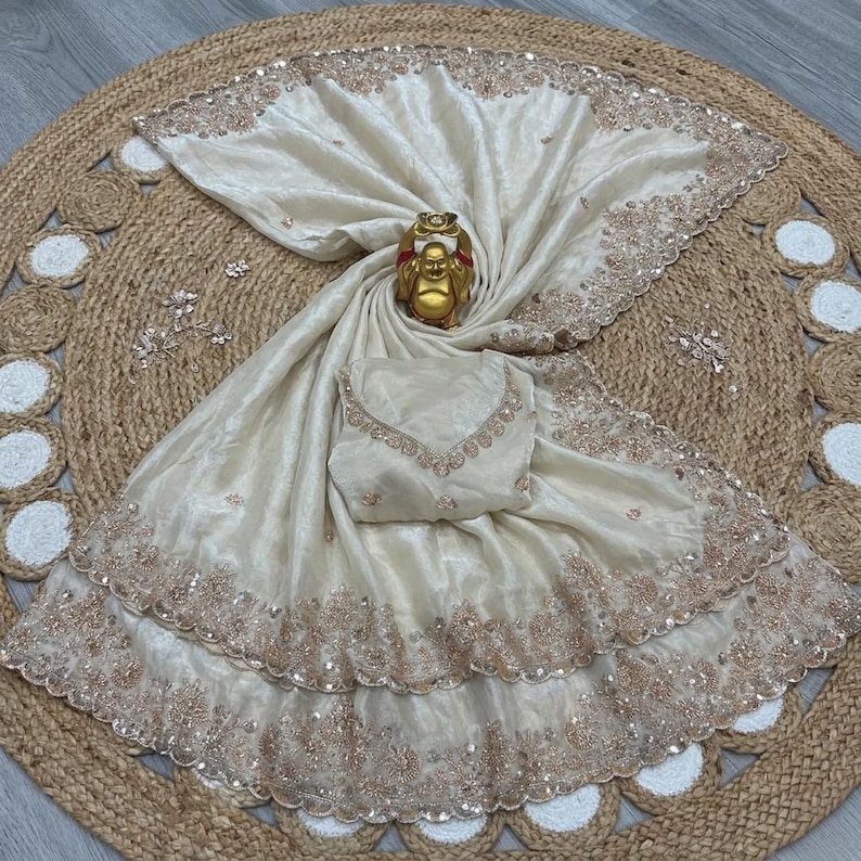 May include: Ivory-colored saree with intricate embroidery and sequin detailing. The saree is draped over a matching blouse and a golden Buddha statue. The ensemble is displayed on a round, woven jute rug.