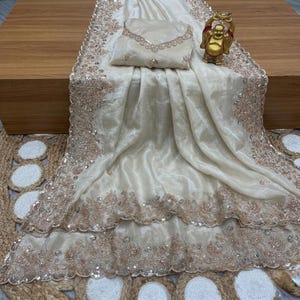 May include: Ivory-colored saree with intricate gold embroidery and sequin detailing. The saree is draped over a wooden surface, with a matching blouse piece and a small gold Buddha statue. The fabric has a shimmering, luxurious appearance.