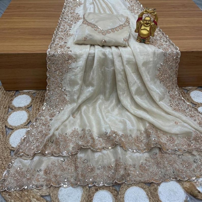 May include: Ivory-colored saree with intricate gold embroidery and sequin detailing. The saree is draped over a wooden surface, with a matching blouse piece and a small gold Buddha statue. The fabric has a shimmering, luxurious appearance.