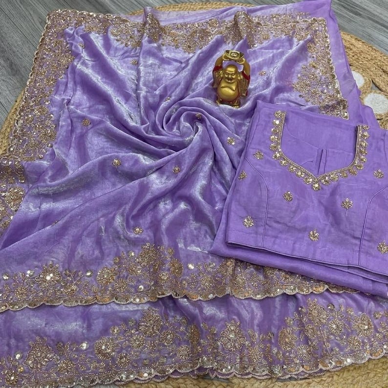 May include: A lavender velvet saree with intricate gold embroidery along the borders and scattered across the fabric. A matching blouse with gold embellishments is placed beside the saree. A small gold Buddha statue sits atop the saree.