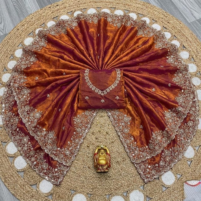 May include: A vibrant orange and burgundy saree with intricate embroidery and embellishments. The saree is fanned out, showcasing its detailed design and a matching blouse. A small gold Buddha statue is at the bottom.