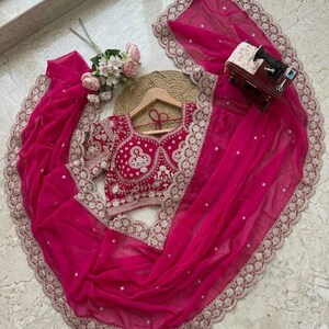 Op de afbeelding: Een levendige fuchsia-kleurige saree-set. De set bevat een geborduurde blouse en een bijpassende dupatta met een delicate kanten rand. De saree is elegant gedrapeerd en toont de vloeiende stof en het ontwerp.