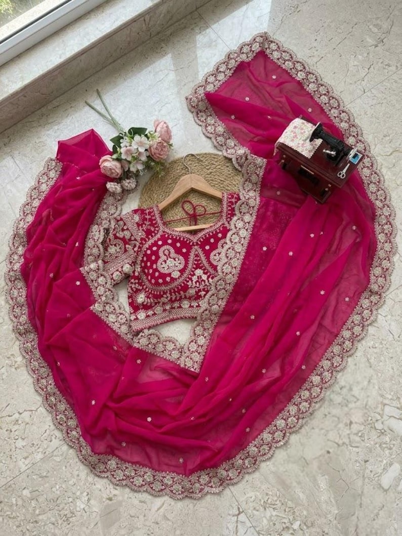 Op de afbeelding: Een levendige fuchsia-kleurige saree-set. De set bevat een geborduurde blouse en een bijpassende dupatta met een delicate kanten rand. De saree is elegant gedrapeerd en toont de vloeiende stof en het ontwerp.