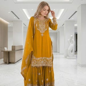 Puede incluir: Traje Sharara bordado amarillo mostaza con dupatta a juego. La t&uacute;nica de manga larga y los pantalones de pernera ancha est&aacute;n adornados con intrincados bordados dorados. El atuendo lo lleva una persona con cabello rubio y aretes dorados.