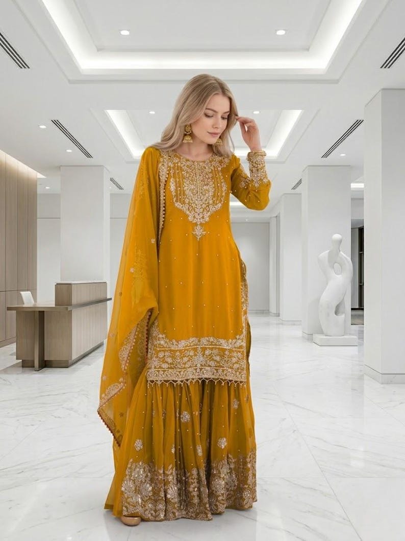 Puede incluir: Traje Sharara bordado amarillo mostaza con dupatta a juego. La t&uacute;nica de manga larga y los pantalones de pernera ancha est&aacute;n adornados con intrincados bordados dorados. El atuendo lo lleva una persona con cabello rubio y aretes dorados.