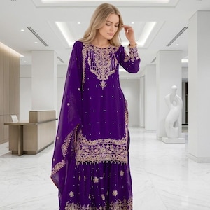 Puede incluir: Un vestido largo y fluido de color púrpura con intrincados bordados dorados. El vestido presenta un cuello redondo, mangas largas y una falda amplia. El bordado dorado se concentra alrededor del cuello, las mangas y el dobladillo. Una bufanda púrpura a juego está drapeada sobre el hombro.