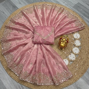 May include: A light pink saree with intricate gold embroidery along the borders and scattered across the fabric. The saree is draped over a matching blouse piece. A gold Buddha statue and decorative coasters are also visible.