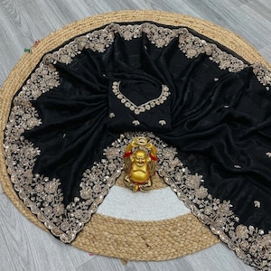 May include: Black saree with intricate gold embroidery along the borders and neckline. The saree is draped over a round, woven mat. A gold Buddha statue is placed in the center.