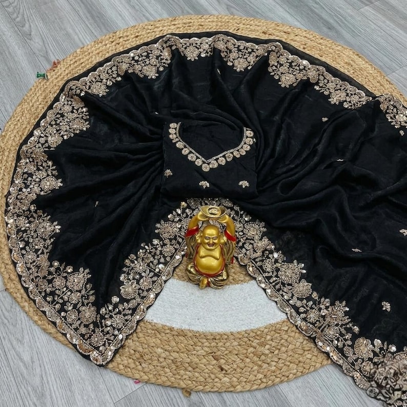 May include: Black saree with intricate gold embroidery along the borders and neckline. The saree is draped over a round, woven mat. A gold Buddha statue is placed in the center.