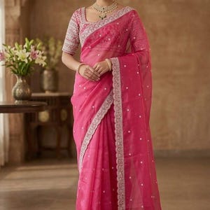 Op de afbeelding: Een felroze sari met ingewikkelde borduursels en een delicate rand. De sari heeft een bijpassende blouse met gedetailleerde mouwen en een vierkante halslijn. De outfit is geaccessoireerd met gouden sieraden, wat een elegante en traditionele look cre&euml;ert.