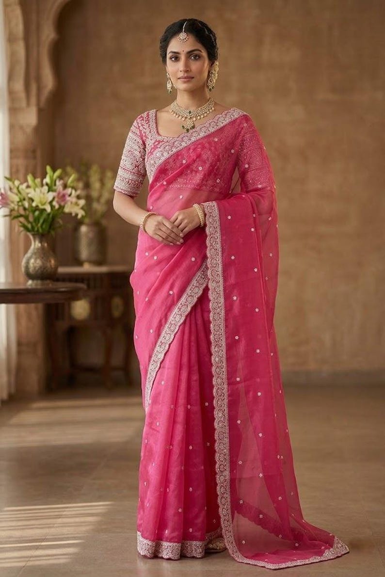 Op de afbeelding: Een felroze sari met ingewikkelde borduursels en een delicate rand. De sari heeft een bijpassende blouse met gedetailleerde mouwen en een vierkante halslijn. De outfit is geaccessoireerd met gouden sieraden, wat een elegante en traditionele look cre&euml;ert.