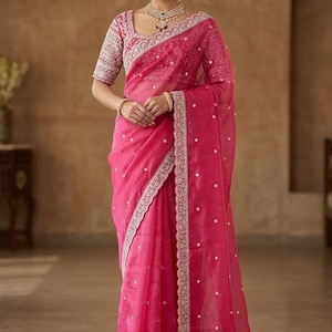 Op de afbeelding: Een felroze sari met ingewikkelde borduursels en een delicate rand. De sari heeft een bijpassende blouse met gedetailleerde mouwen en een vierkante halslijn. De outfit is geaccentueerd met gouden sieraden, wat een elegante en traditionele uitstraling cre&euml;ert.