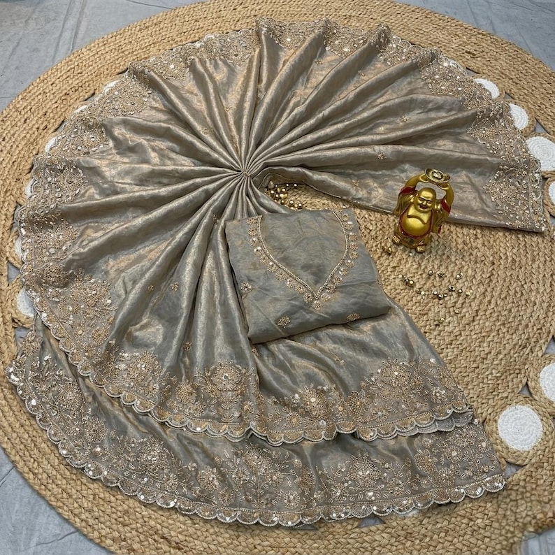 May include: A beige silk saree with intricate gold embroidery and sequin detailing. The saree is draped over a matching blouse and a decorative gold Buddha statue. The fabric has a subtle sheen, and the design is ornate.