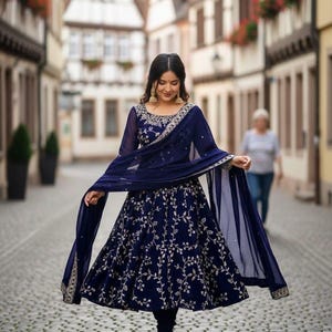 Peut inclure: Une robe Anarkali brodée bleu marine avec un dupatta assorti. La robe présente des broderies florales complexes dans une couleur plus claire. La modèle porte des boucles d'oreilles et des chaussures dorées.
