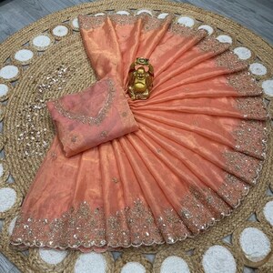 May include: Peach-colored saree with intricate embroidery along the border and a matching blouse. A golden Buddha statue sits atop the folded fabric. The saree is displayed on a woven jute rug.