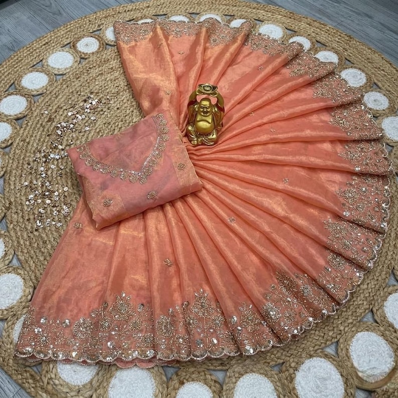 May include: Peach-colored saree with intricate embroidery along the border and a matching blouse. A golden Buddha statue sits atop the folded fabric. The saree is displayed on a woven jute rug.