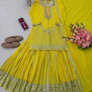 Pu&ograve; includere: Un vivace completo sharara giallo con un dupatta abbinato, con ricami dettagliati e abbellimenti di paillettes. La parte superiore presenta uno scollo rosa e argento. I pantaloni a gamba larga e il dupatta sono anch'essi decorati con dettagli argentati. Sono presenti anche scarpe ricamate e un bouquet di fiori rosa.