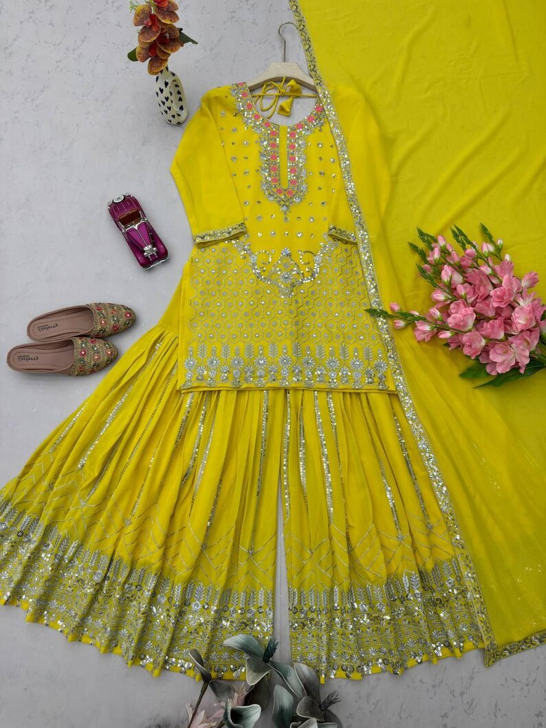 Pu&ograve; includere: Un vivace completo sharara giallo con un dupatta abbinato, con ricami dettagliati e abbellimenti di paillettes. La parte superiore presenta uno scollo rosa e argento. I pantaloni a gamba larga e il dupatta sono anch'essi decorati con dettagli argentati. Sono presenti anche scarpe ricamate e un bouquet di fiori rosa.