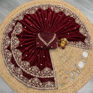 May include: A maroon velvet saree with intricate gold embroidery and embellishments. The saree is draped over a matching blouse, with a gold figurine and decorative elements on a woven jute mat.
