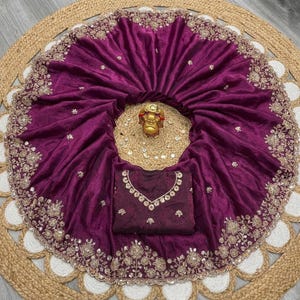 May include: A deep purple sari with intricate gold embroidery along the border and scattered across the fabric. The sari is draped over a matching blouse with detailed neckline embellishments. A small gold statue sits in the center.