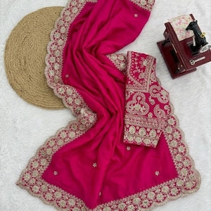 Op de afbeelding: Een levendige fuchsia sari met ingewikkelde gouden borduursels langs de randen en een bijpassende blouse. De sari is elegant gedrapeerd en toont het gedetailleerde vakmanschap en ontwerp. Het is een traditioneel kledingstuk.