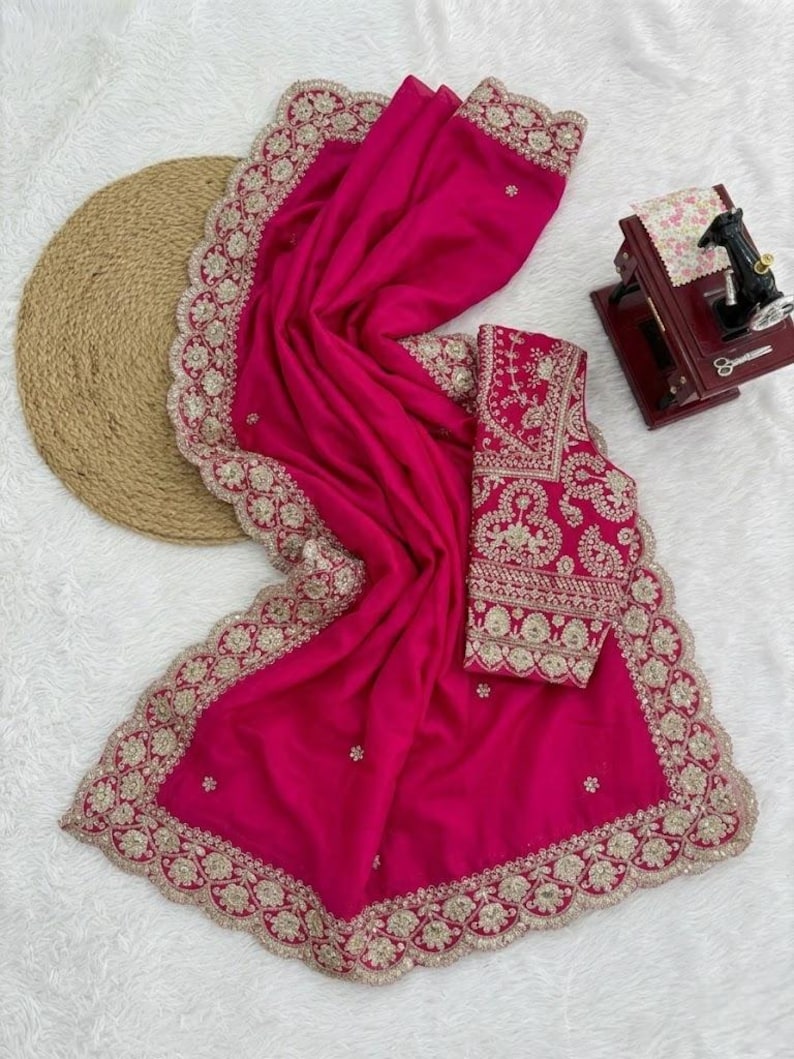 Op de afbeelding: Een levendige fuchsia sari met ingewikkelde gouden borduursels langs de randen en een bijpassende blouse. De sari is elegant gedrapeerd en toont het gedetailleerde vakmanschap en ontwerp. Het is een traditioneel kledingstuk.