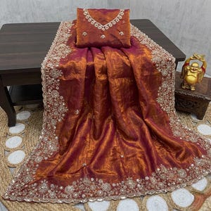 May include: A luxurious, shimmering saree in shades of burgundy and gold, with intricate embroidery along the borders and neckline. The fabric drapes elegantly, showcasing a rich, textured appearance. A matching blouse piece is included.