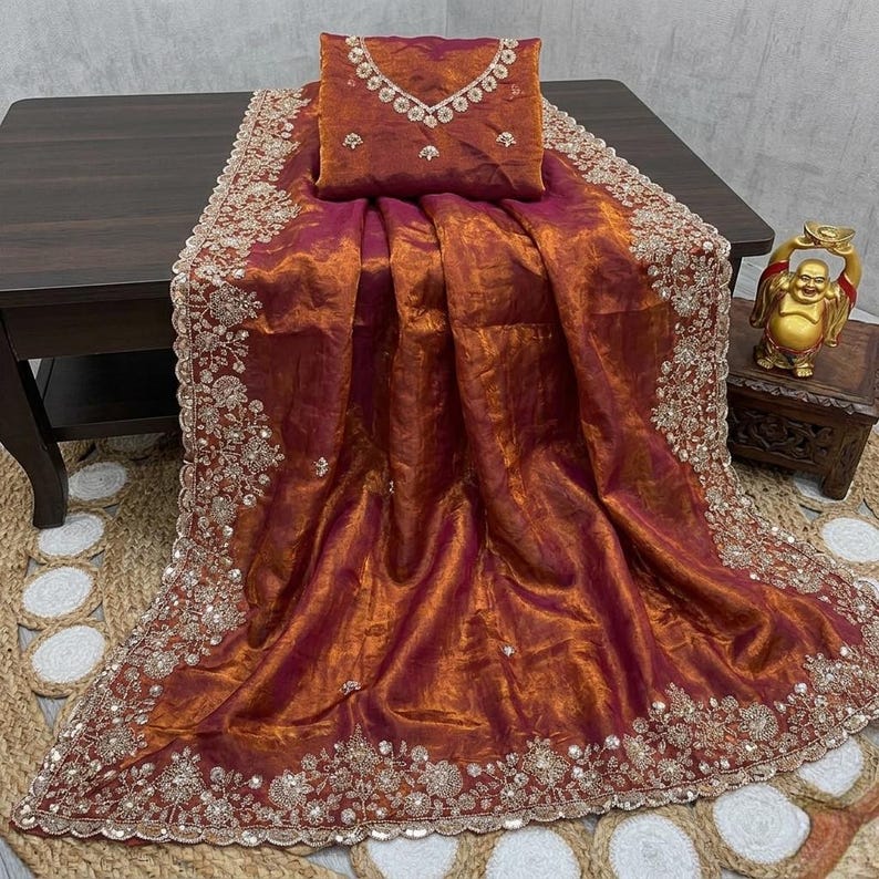 May include: A luxurious, shimmering saree in shades of burgundy and gold, with intricate embroidery along the borders and neckline. The fabric drapes elegantly, showcasing a rich, textured appearance. A matching blouse piece is included.