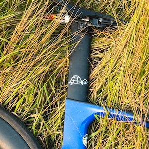 Könnte beinhalten: Nahaufnahme eines blau-schwarzen Fahrradrahmens mit einem weißen Schildkrötenlogo auf der Sattelstütze. Das Fahrrad ist teilweise sichtbar und liegt in hohem, trockenem Gras. Der Rahmen ist leuchtend blau.