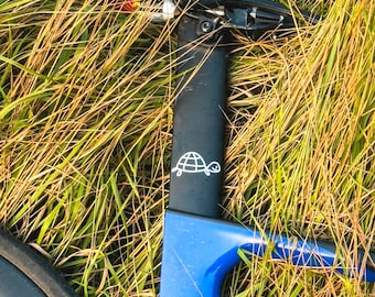 Könnte beinhalten: Nahaufnahme eines blau-schwarzen Fahrradrahmens mit einem weißen Schildkrötenlogo auf der Sattelstütze. Das Fahrrad ist teilweise sichtbar und liegt in hohem, trockenem Gras. Der Rahmen ist leuchtend blau.