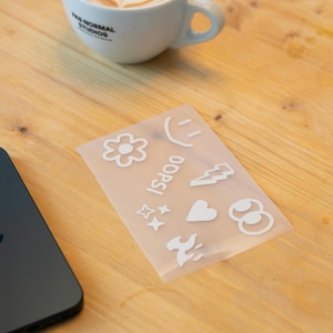 Könnte beinhalten: Ein transparentes Stickerbogen mit weißen Grafiken, darunter eine Blume, ein Smiley, ein Blitz, Sterne, ein Herz und Flammen. Das Wort "OOPS!" ist vertikal aufgedruckt. Eine Kaffeetasse und ein Smartphone sind ebenfalls zu sehen.