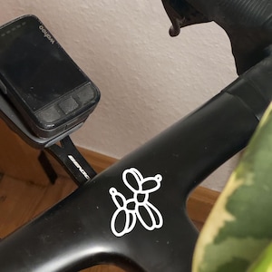 May include: A black bicycle frame with a white balloon dog sticker. A Wahoo cycling computer is mounted on the handlebars. The bike is near a green plant. The frame is made of carbon fibre.