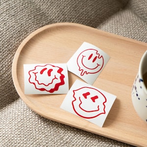 Peut inclure: Trois autocollants blancs avec des motifs de visages souriants rouges et déformés sont présentés sur un plateau en bois. Les autocollants ont des expressions différentes, dont un visage qui fond et un visage légèrement en colère. Une tasse avec des mouchetures bleues est visible.