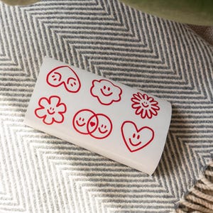 Könnte beinhalten: Ein weißes Stickerbogen mit roten, strichgezeichneten Smiley-Designs. Das Bogen zeigt verschiedene Formen, darunter Herzen, Blumen und Wolken, jeweils mit einem lächelnden Gesicht. Das Stickerbogen liegt auf einer grau-weiß gemusterten Decke.