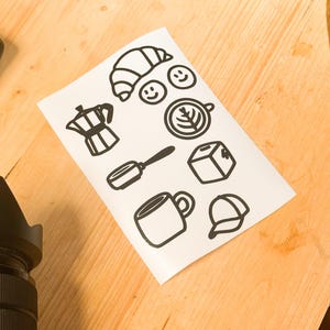 May include: A white sheet featuring black line drawings of coffee-themed icons. These include a croissant, smiley faces, a coffee pot, a spoon, a cube, a mug, a cap, and a coffee cup with a leaf design.