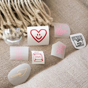 Könnte beinhalten: Eine Sammlung von Aufklebern mit verschiedenen Designs, darunter ein rotes Herz mit Smiley-Gesicht, eine Kaffeemaschine, eine Blume und der Text "LOST Ctrl". Die Aufkleber haben verschiedene Formen und Größen mit rosa und roten Akzenten.