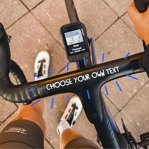 Może przedstawiać: Czarna kierownica rowerowa z białym napisem CHOOSE YOUR OWN TEXT oraz zamontowanym centralnie licznikiem rowerowym Wahoo wyświetlającym dane z trasy. Kierownica owinięta jest czarną taśmą, a niebieskie linie graficzne podkreślają napis.