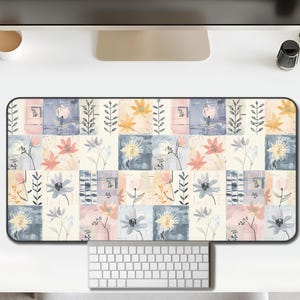 May include: Rectangular desk mat with a floral pattern. The design features a patchwork of squares with watercolor-style flowers in shades of pink, blue, yellow, and cream. Black border surrounds the mat. A keyboard and mouse are visible.