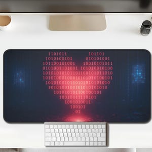 Op de afbeelding: Een rechthoekige bureaumat met een hart van binaire code in rood op een donkerblauwe achtergrond. Een wit toetsenbord en muis zijn zichtbaar. De mat ligt op een witte tafel, samen met een computermonitor en kantoorbenodigdheden.