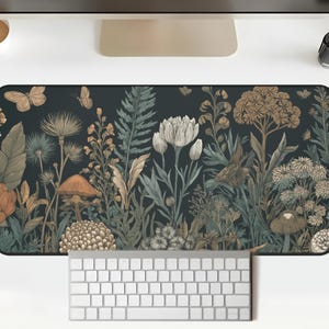 Puede incluir: Alfombrilla de escritorio rectangular con una ilustración botánica de setas, flores y mariposas en tonos verdes, marrones y beige. Un teclado blanco está colocado en la parte inferior.