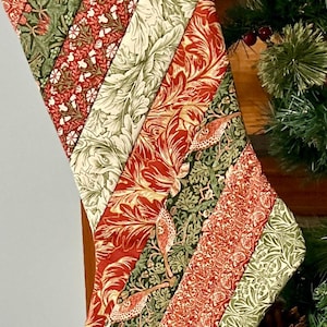 May include: Quilted Christmas stocking with a patchwork design. The fabric features a variety of patterns in red, green, and cream, including floral and bird motifs. The stocking is displayed near a Christmas tree.