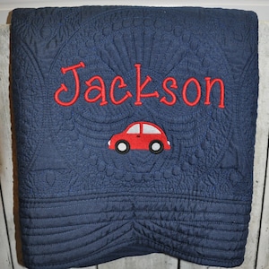 Puede incluir: Una manta acolchada de color azul marino con intrincados patrones de costura en toda la superficie. El centro muestra el nombre Jackson bordado en hilo rojo brillante, situado sobre un peque&ntilde;o gr&aacute;fico de coche rojo con detalles en blanco y negro.