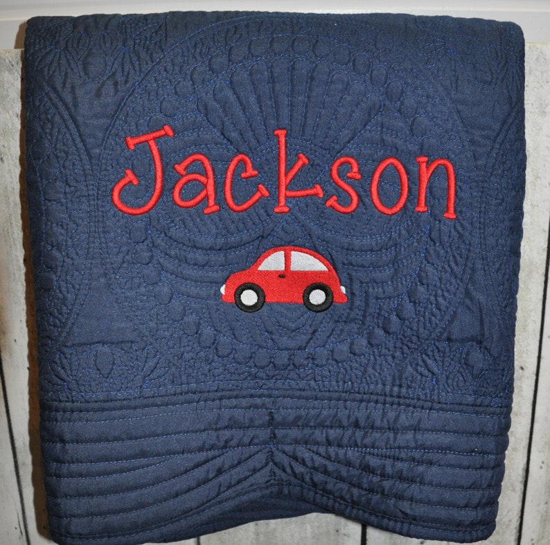 Puede incluir: Una manta acolchada de color azul marino con intrincados patrones de costura en toda la superficie. El centro muestra el nombre Jackson bordado en hilo rojo brillante, situado sobre un peque&ntilde;o gr&aacute;fico de coche rojo con detalles en blanco y negro.