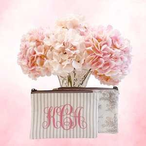 May include: Two canvas zipper pouches with a floral arrangement in the background. One pouch has a gray and white striped pattern with a pink monogram. The other pouch has a gray and white toile pattern.