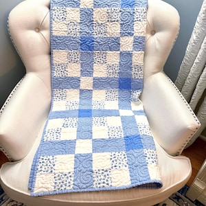 May include: A rectangular quilt featuring a grid pattern of alternating squares in shades of light blue, medium blue, and white. The fabric squares display various textures, including solid colors, small floral prints, and intricate swirling stitch patterns.