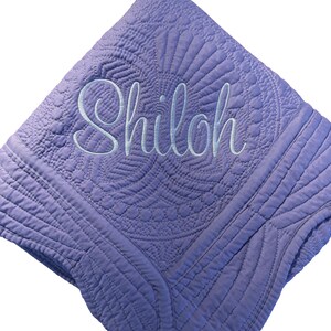 Puede incluir: Una manta acolchada de color lavanda con intrincados patrones de costura y el nombre Shiloh bordado en una fuente cursiva azul claro en el centro. La tela tiene una superficie texturizada con detalles geom&eacute;tricos y florales.
