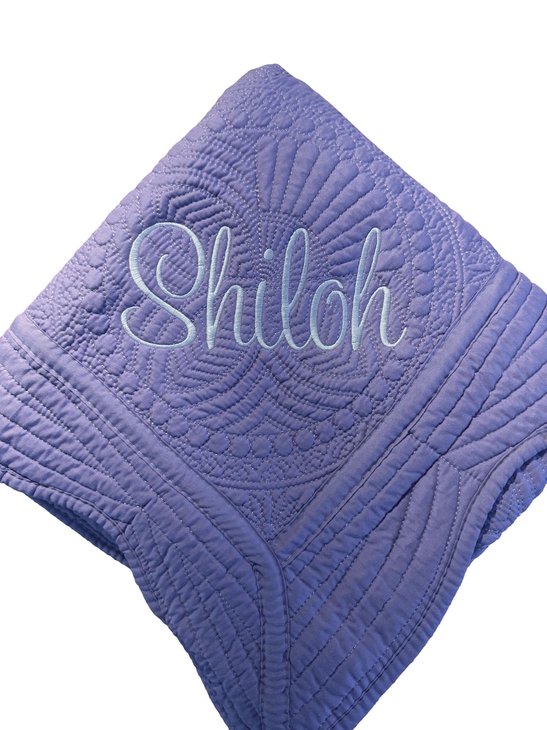 Puede incluir: Una manta acolchada de color lavanda con intrincados patrones de costura y el nombre Shiloh bordado en una fuente cursiva azul claro en el centro. La tela tiene una superficie texturizada con detalles geom&eacute;tricos y florales.