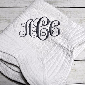 Puede incluir: Una manta acolchada blanca con un monograma bordado en gris oscuro con las letras ACC en letra cursiva. La tela presenta un patr&oacute;n de costuras texturizado en toda la superficie y la manta est&aacute; doblada en forma cuadrada.