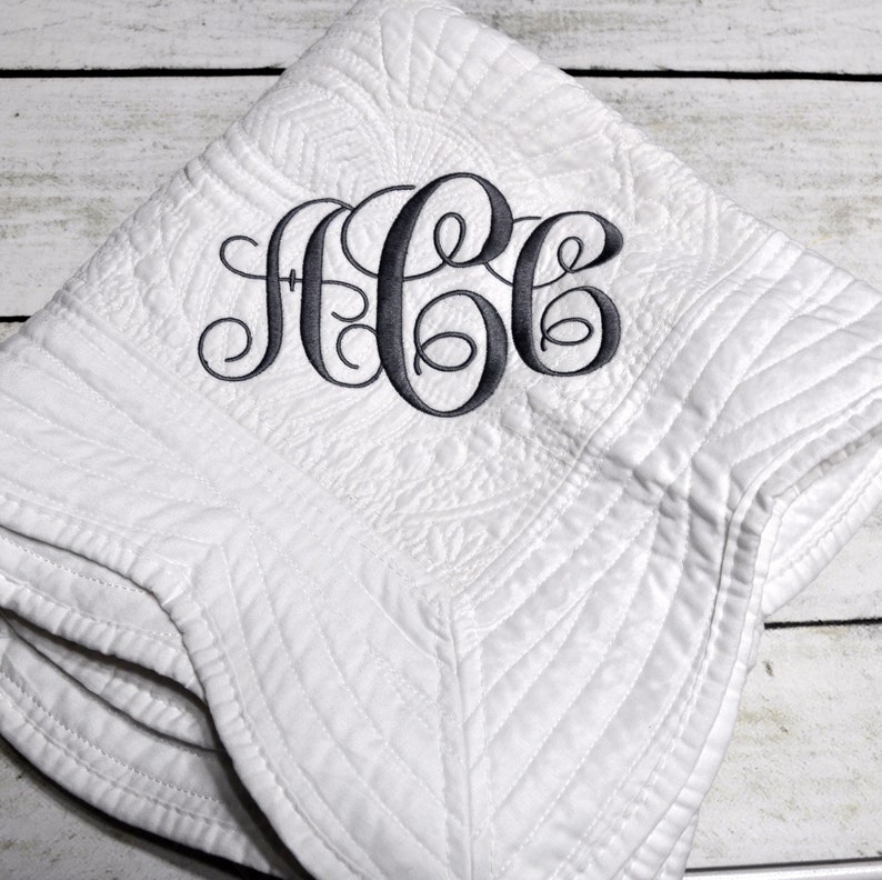 Puede incluir: Una manta acolchada blanca con un monograma bordado en gris oscuro con las letras ACC en letra cursiva. La tela presenta un patr&oacute;n de costuras texturizado en toda la superficie y la manta est&aacute; doblada en forma cuadrada.